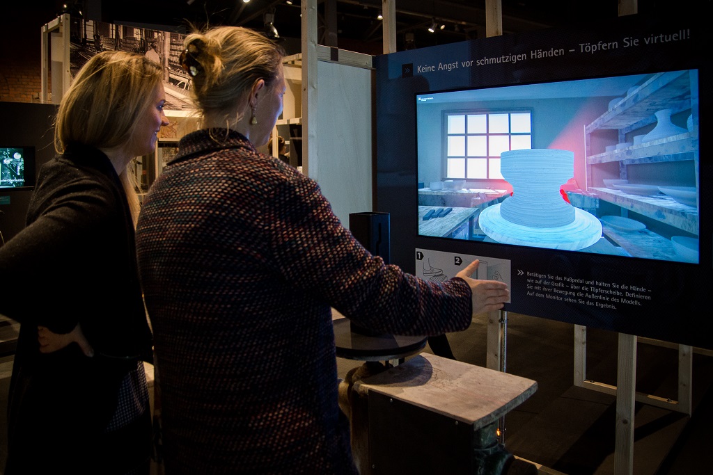 Frau steht vor einem Sensor und einem Bildschirm. Die modellierenden Bewegungen ihrer Hände um den Sensor herum lassen auf dem Bildschirm ein virutelles Gefäß Gestalt annehmen.